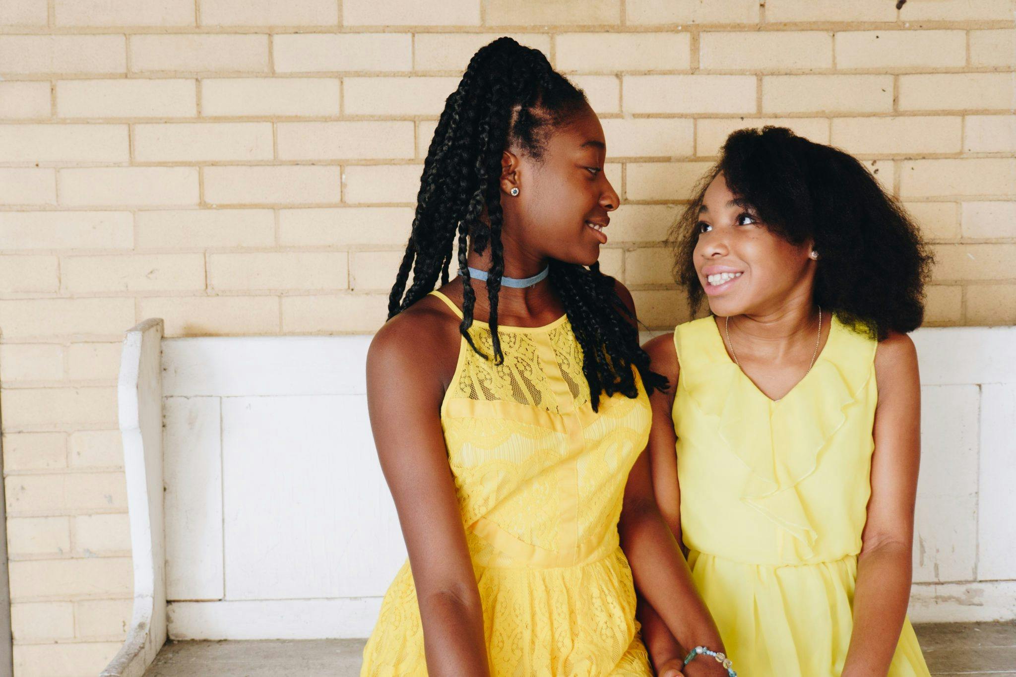 Two sisters in yellow dresses smiling at each other, radiating joy indoors.